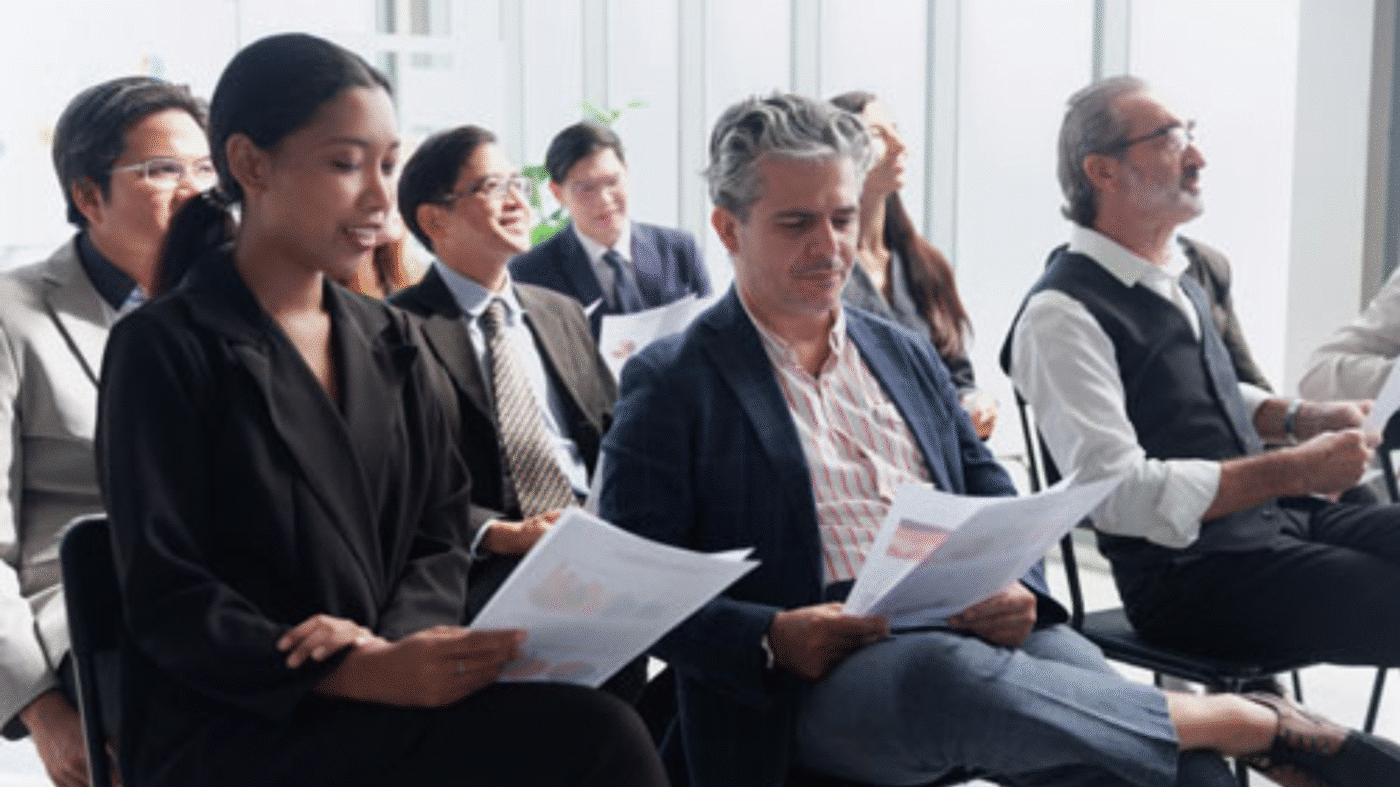 A diverse group of people in business attire sit in chairs, holding papers and attentively listening during a meeting or seminar in a bright, modern office space.