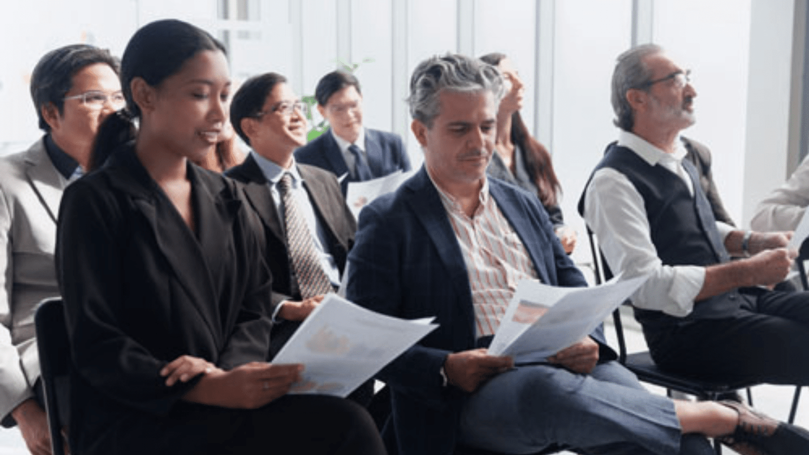 A diverse group of people in business attire sit in chairs, holding papers and attentively listening during a meeting or seminar in a bright, modern office space.