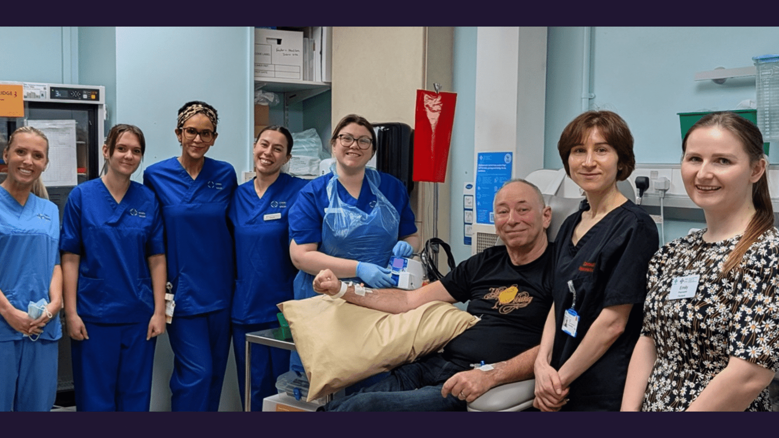 A group of healthcare professionals, some in blue scrubs and others in casual work attire, gather and smile around a seated patient receiving treatment in a clinical room.