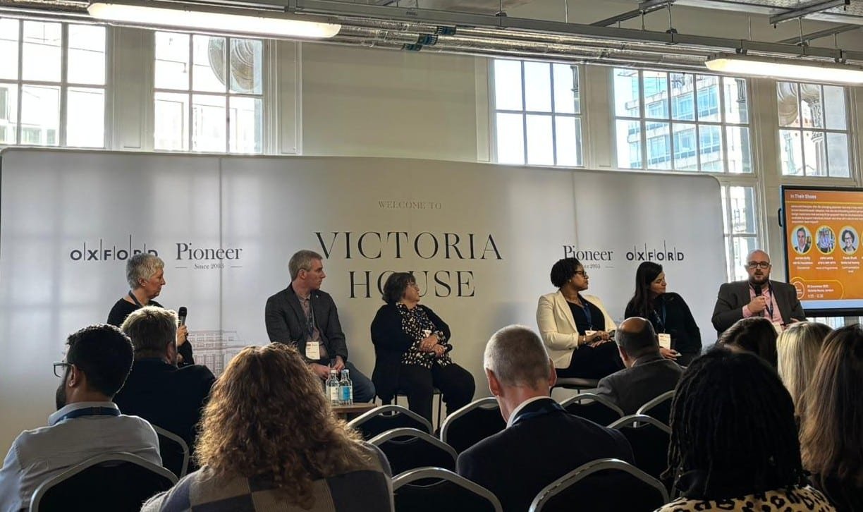 A panel of five people sits on stage at a conference in front of an audience. Behind them is a backdrop with Victoria House, Oxford, and Pioneer logos. A presenter holds a microphone, and a screen displays information on the right-hand side.