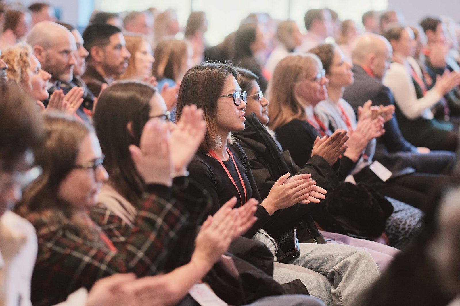 A diverse group of people sitting in rows and applauding at an indoor event or conference, with most audience members facing forwards and appearing attentive.