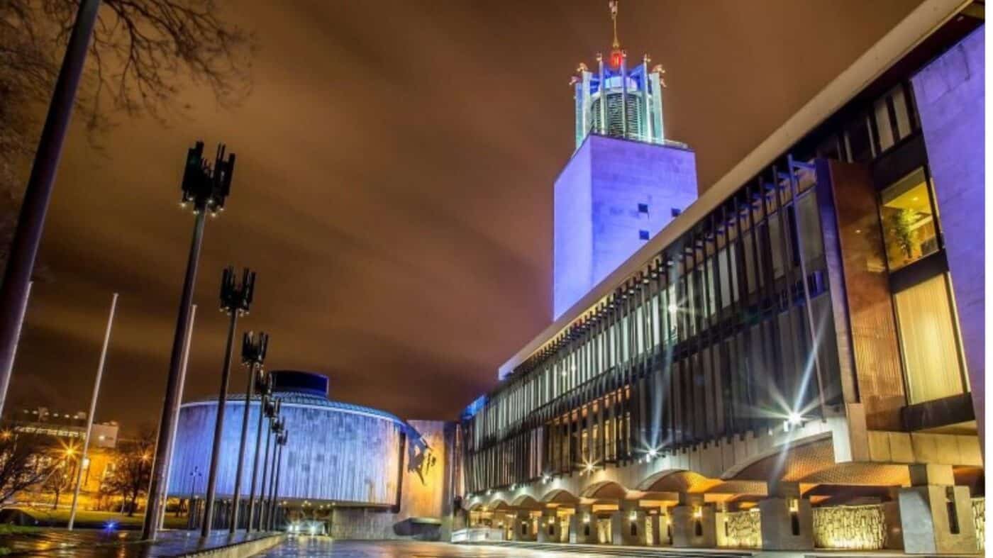 A modern, illuminated building with a tall tower and glass facade at night, with dramatic clouds and streetlights, and an empty square in the foreground.
