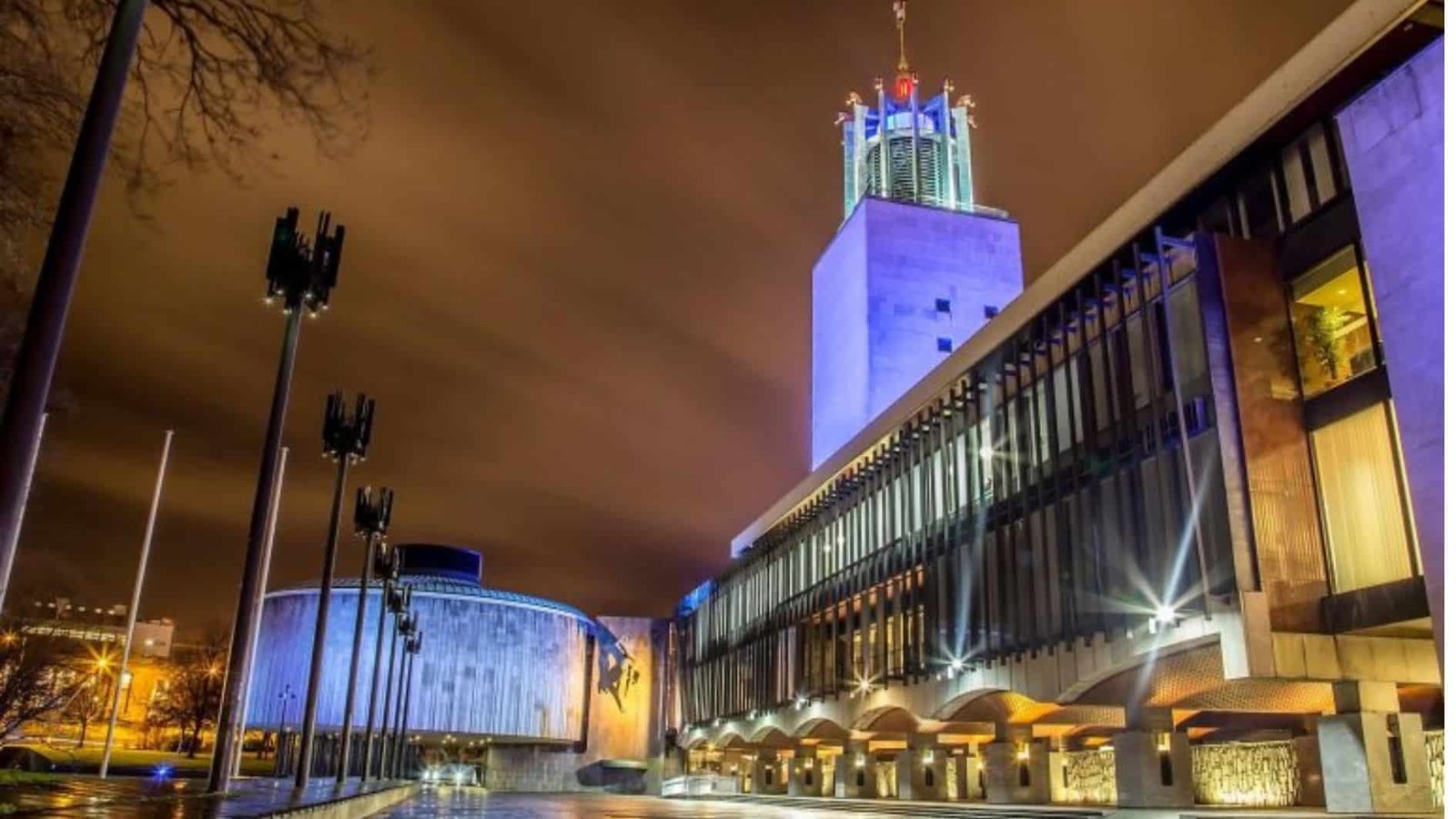 A modern, illuminated building with a tall tower and glass facade at night, with dramatic clouds and streetlights, and an empty square in the foreground.