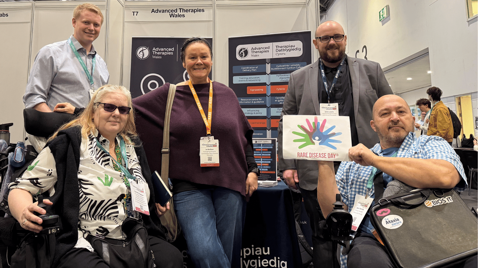 Five people smile at a conference stand for Advanced Therapies Wales. Two people use wheelchairs, and one holds a sign that says Rare Disease Day with a colourful handprint logo.