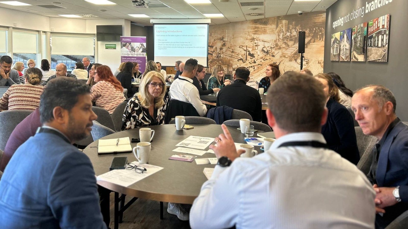 A group of people sit around circular tables in a conference room, engaged in discussion. Papers, mugs, and notebooks are on the tables. A presentation screen and informational banners are visible in the background.