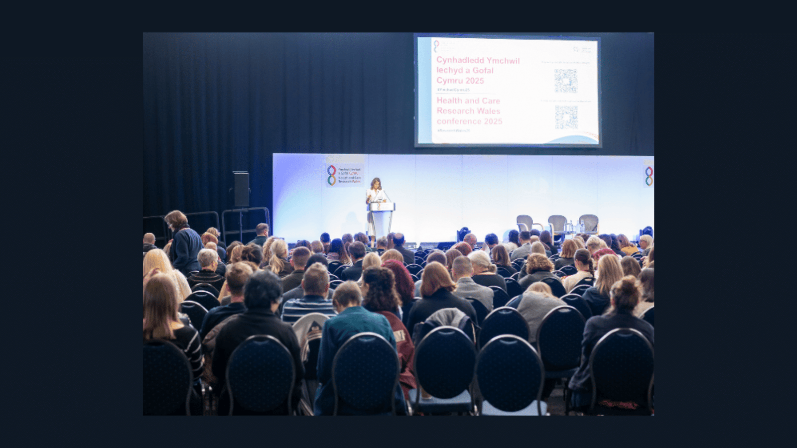 A speaker addresses a seated audience at a conference. A large screen behind shows event details in English and Welsh, titled Health and Care Research Wales conference 2023.